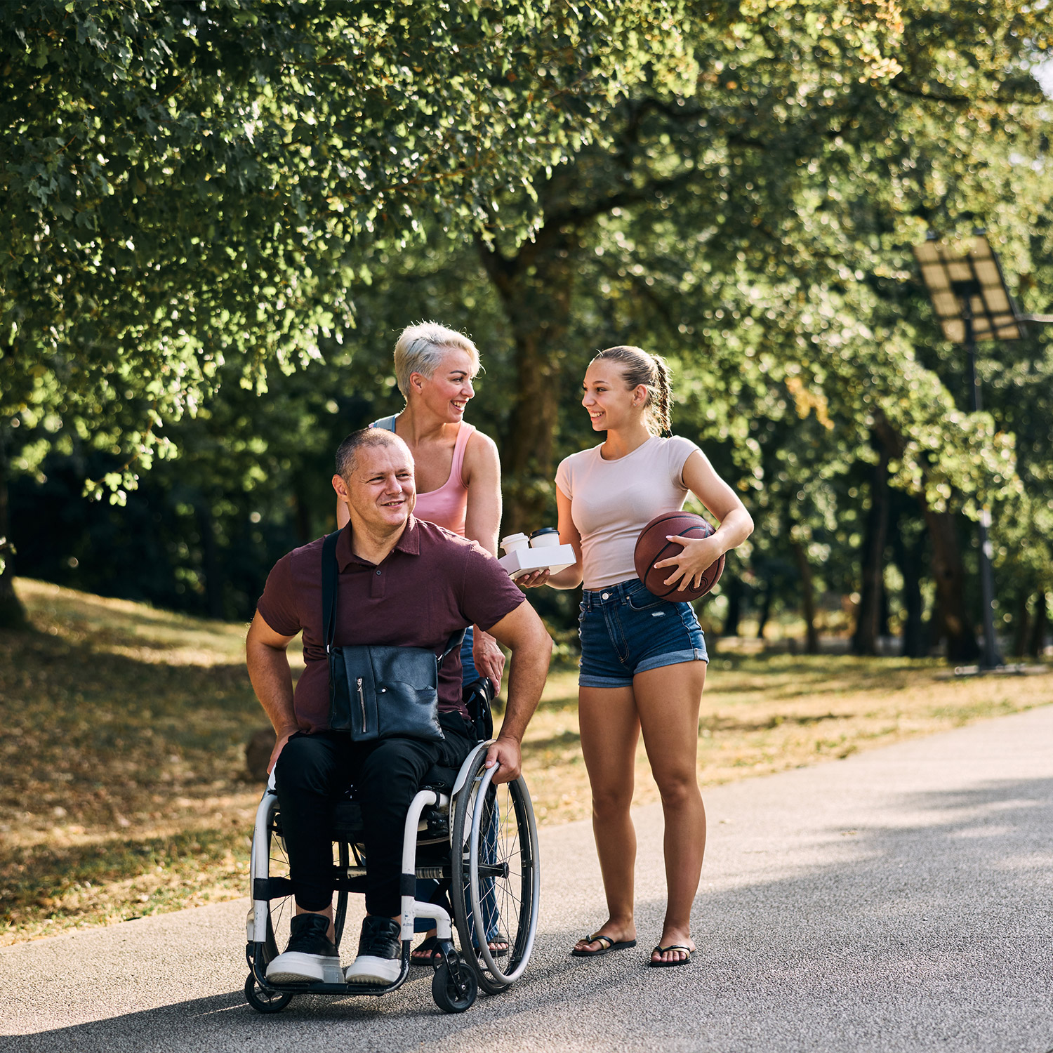Zwei Frauen sind gemeinsam im Park unterwegs mit einem Rollstuhlfahrer